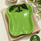Green ceramic pepper-shaped dish on a beige cloth with a bowl of nuts in the background.