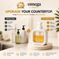Omega Shop countertop dispenser set shown in a kitchen-like counter scene with a sink, bottles, and a large featured white pump dispenser filled with yellow liquid on a marble base. Text overlays compare the old messy countertop way with the new Omega Way style, and the Omega Shop logo appears at the top.